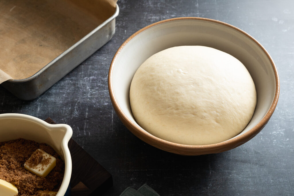 risen dough in a mixing bowl, a baking pan lined with parchment, and brown sugar and butter in a small saucepan