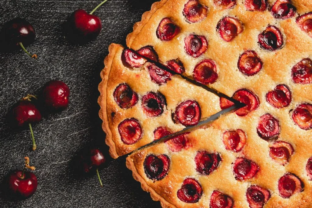 Overhead landscape view of a baked cherry tart with a cut slice and whole cherries scattered on dark surface