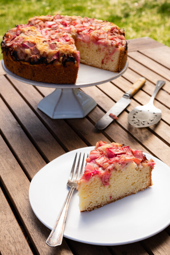 a slice of rhubarb cake on a white plate, rhubarb cake on a cake stand on a wooden table