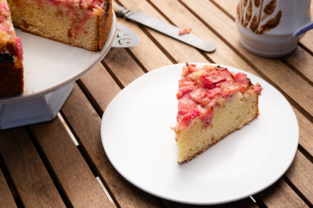a slice of rhubarb cake on a white plate with a coffee mug, knife, and cake stand