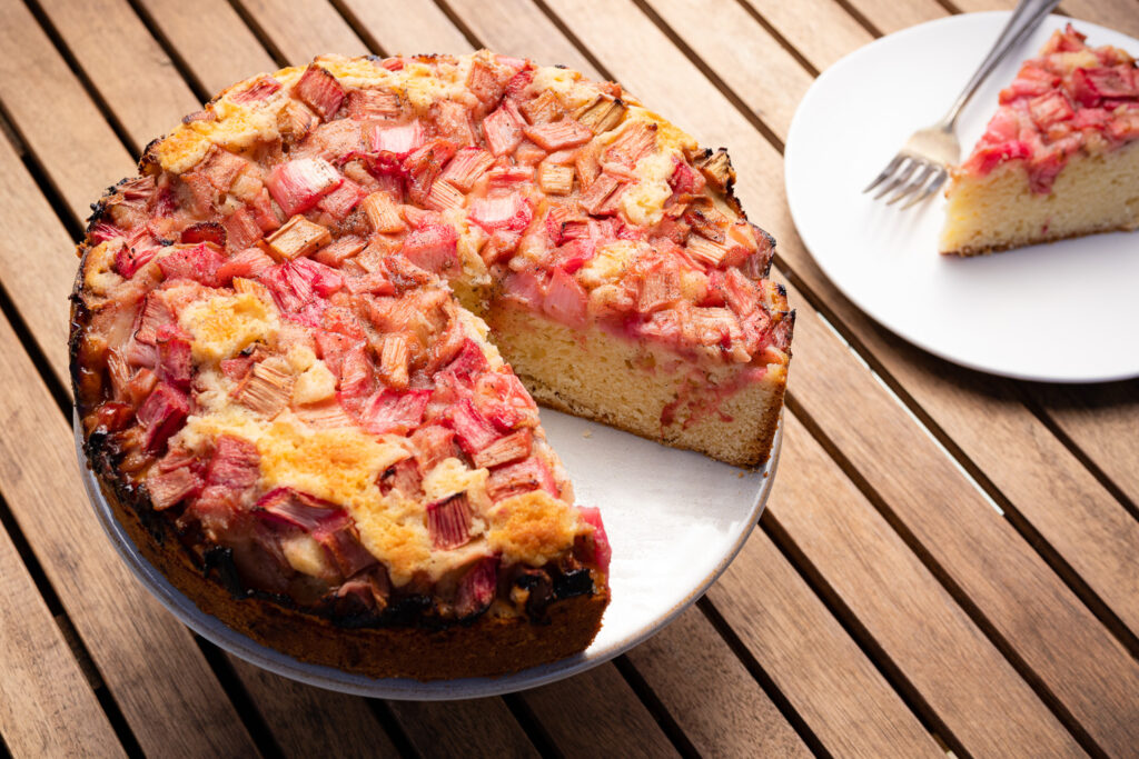 rhubarb cake on a cake stand and a slice of cake on a white plate on a wood table