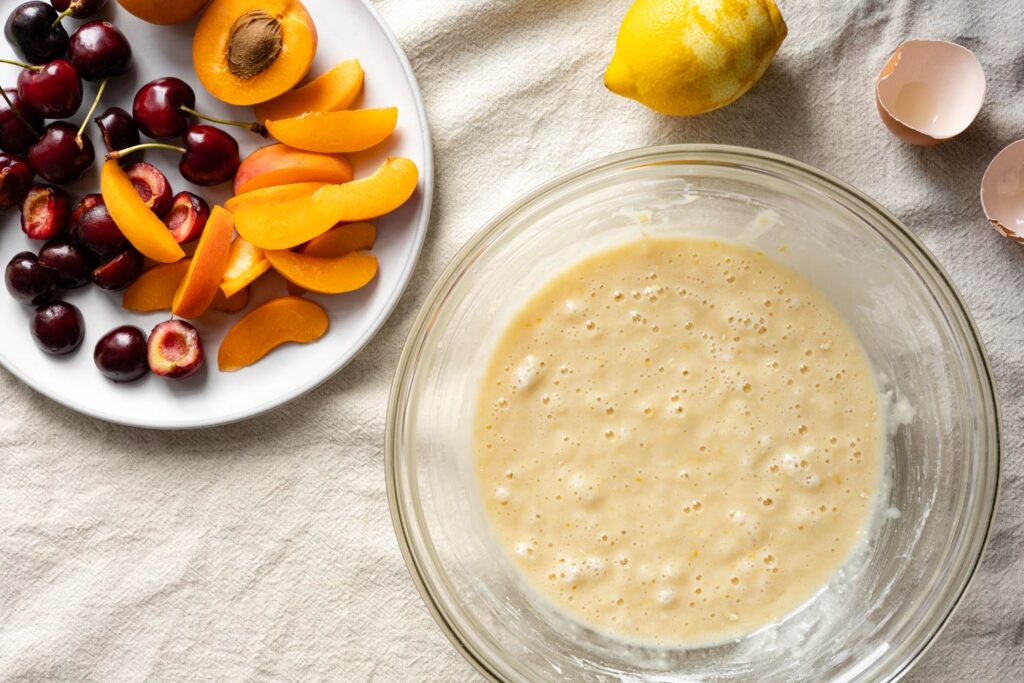 bublanina cake batter in a mixing bowl, cherries and apricots on a white plate, a lemon, and egg shells on a linen tablecloth