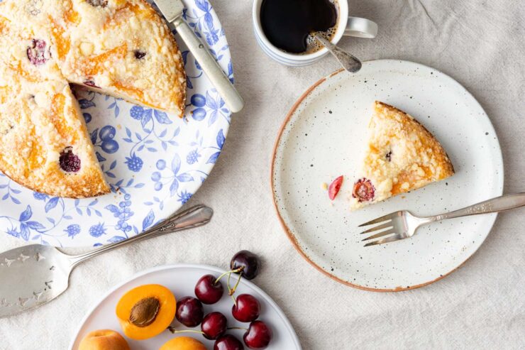 bublanina cake on a blue floral plate, silver-plated cake server, a plate of cherries and apricots, a cup of coffee, and a slice of cake