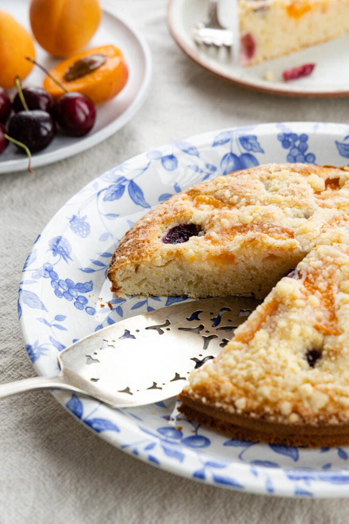 bublanina cake on a blue floral plate, silver-plated cake server, a plate of cherries and apricots, and a slice of cake