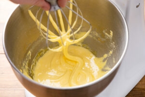 Pale yellow whipped egg yolks clinging to a stand mixer whisk, showing a thick ribbon consistency