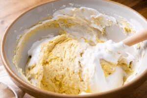 White egg foam being folded into golden batter with a spatula, showing two-tone streaks in the bowl