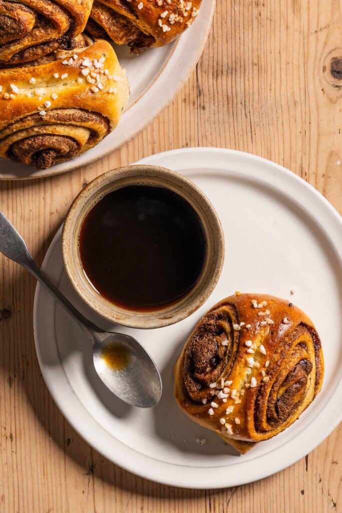 A cinnamon bun, a small cup of coffee, and a coffee spoon on a small ceramic plate next to a larger plate with a stack of cinnamon buns