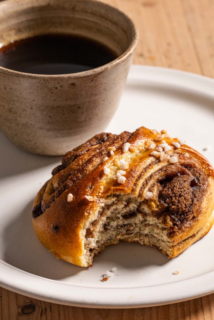 A close up of a korvapuusti with a bite taken out of it in front of a cup of coffee on a ceramic plate