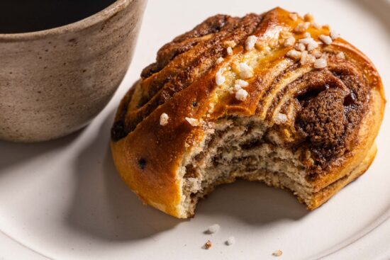 A close up of a korvapuusti with a bite taken out of it in front of a cup of coffee on a ceramic plate