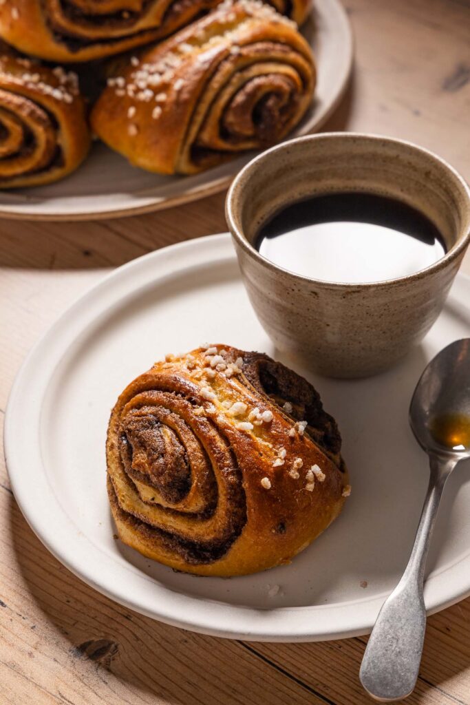 A cinnamon bun, a small cup of coffee, and a coffee spoon on a small ceramic plate next to a larger plate with a stack of cinnamon buns