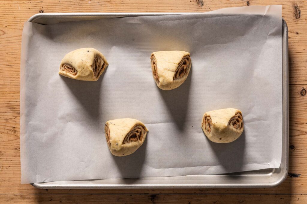 An overhead photo of four unbaked cinnamon buns with the wide side down and the narrow side facing up on a baking sheet lined with parchment paper