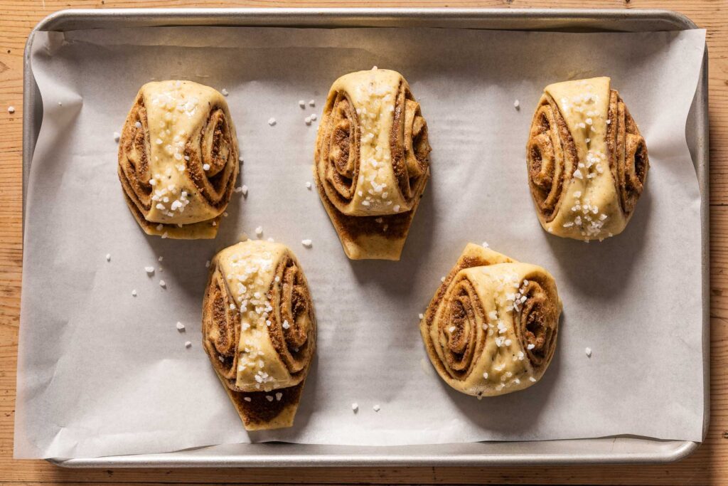 An overhead photo of the cinnamon buns after proofing topped with egg wash and pearl sugar on a parchment paper lined baking sheet