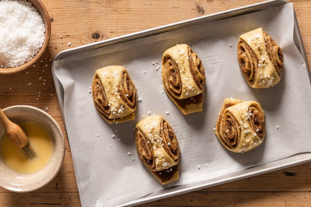 An overhead photo of the cinnamon buns after proofing topped with egg wash and pearl sugar on a parchment paper lined baking sheet next to a bowl of egg wash and a bowl of extra pearl sugar