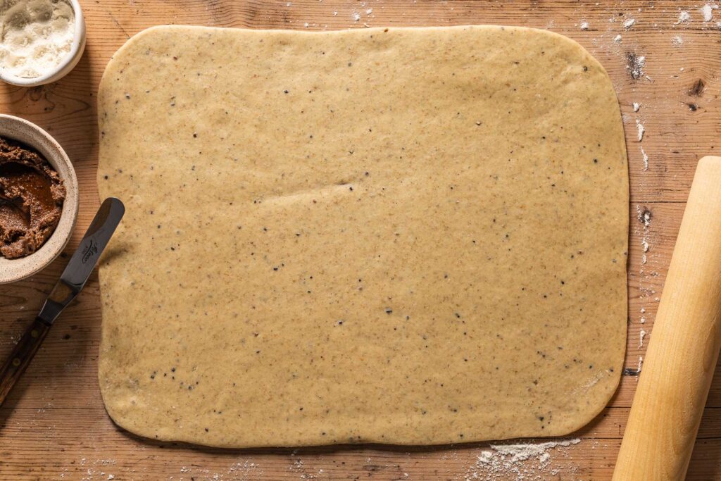 An overhead photo of the dough rolled out into a rectangle on the counter. Next to the dough is a bowl of flour, a bowl with the cinnamon filling, an offset spatula, and a wooden rolling pin