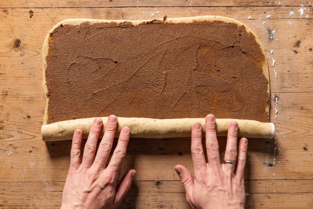 An overhead photo of two hands rolling the cinnamon bun dough into a log