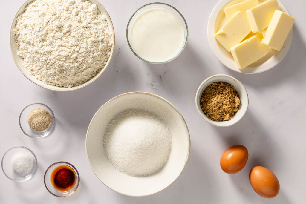 Flour, milk, butter, granulated sugar, brown sugar, eggs, instant yeast, vanilla extract, and salt on a marble surface